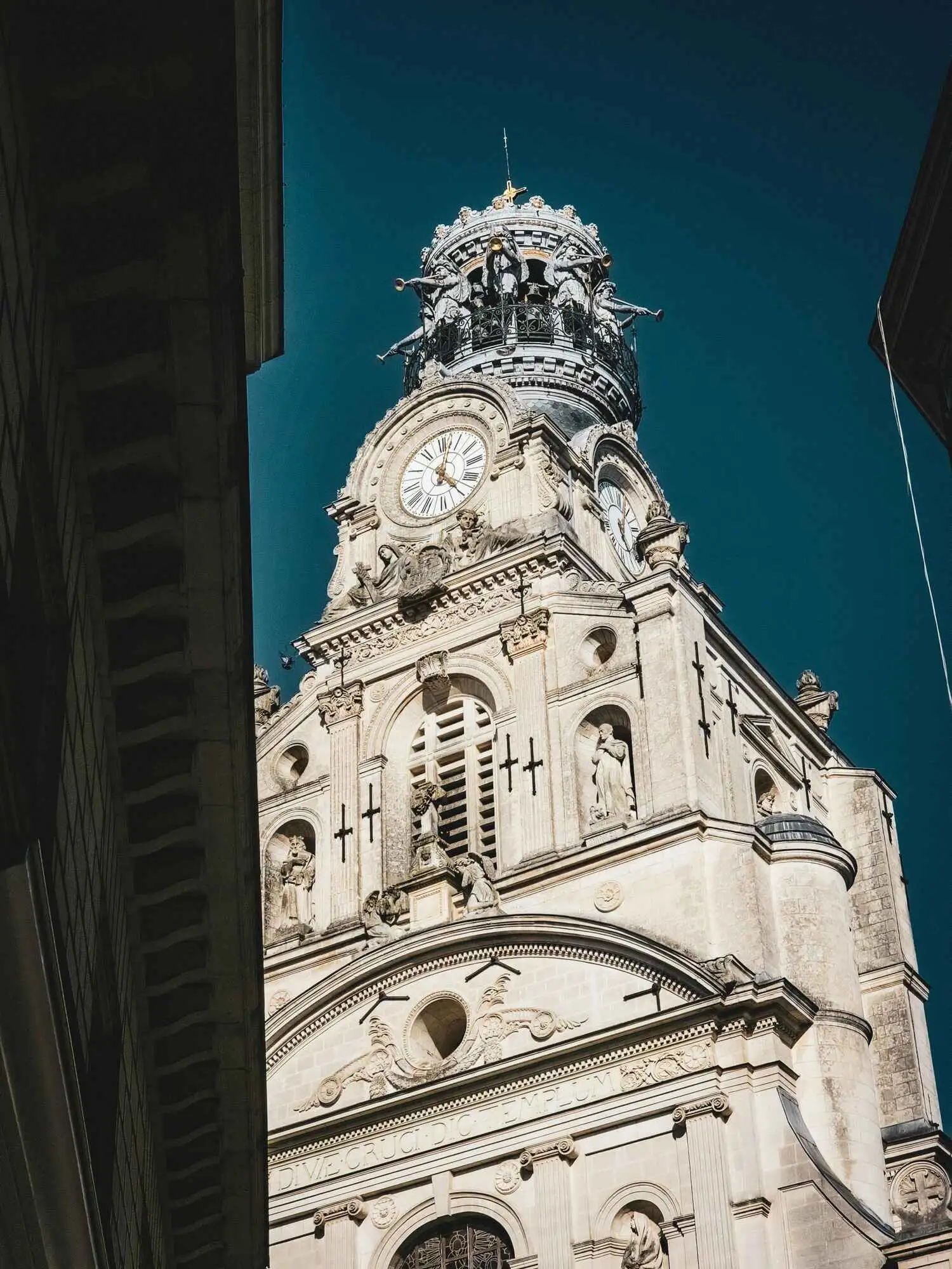 Vue de la tour de l'église Sainte-Croix à Nantes, avec ses statues et son horloge sous un ciel bleu.