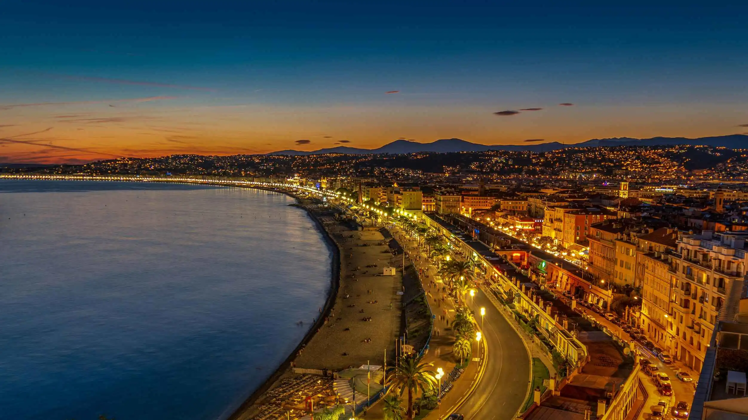 Vue nocturne de la Promenade des Anglais à Nice, avec ses lumières scintillantes, la plage et la mer Méditerranée sous un ciel au coucher du soleil.